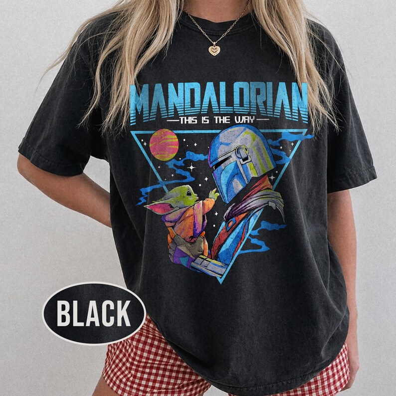May include: Black t-shirt with a graphic of The Mandalorian and Grogu, and the text "MANDALORIAN - THIS IS THE WAY" in blue. The design includes a planet and a triangular frame. The shirt is a dark colour.