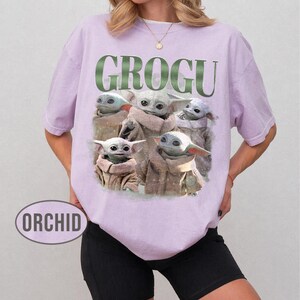 May include: A light purple t-shirt featuring the word "GROGU" in green block letters above a graphic of Grogu from The Mandalorian. The graphic shows Grogu in various poses. The word "ORCHID" is printed on the bottom left.