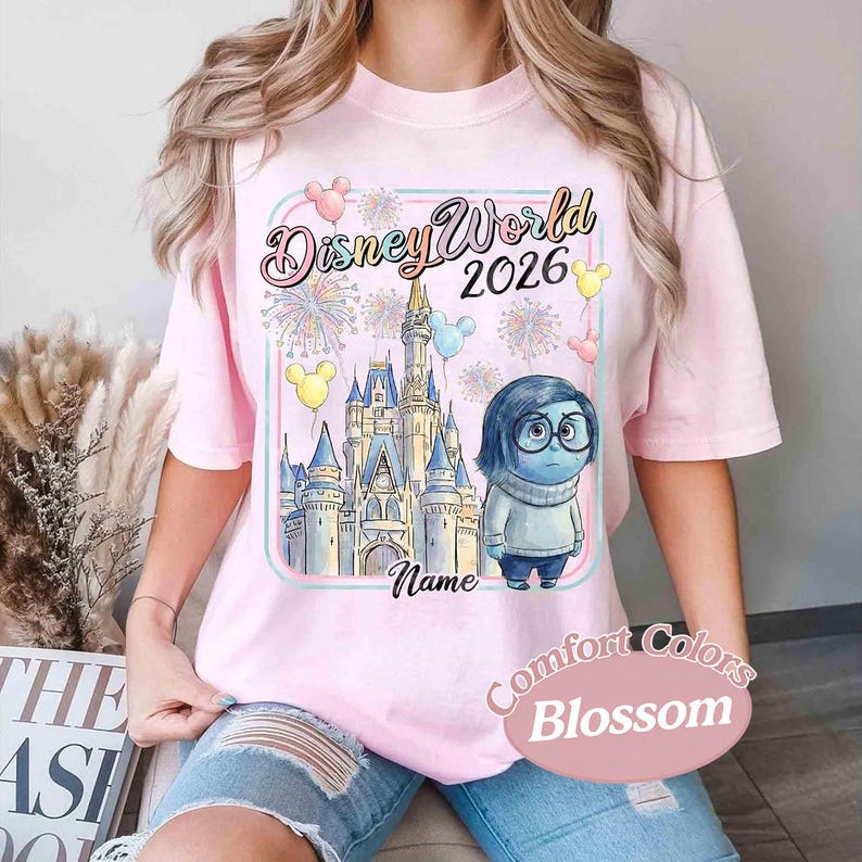 May include: Pink t-shirt with a Disney World 2026 graphic. The design includes a castle, fireworks, balloons, and a cartoon character. The shirt is labeled "Comfort Colors Blossom".