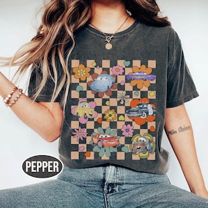 May include: Dark grey t-shirt with a retro design of cartoon cars and flowers on a chequered background. The word "PEPPER" is printed at the bottom. The shirt has short sleeves and a crew neck.