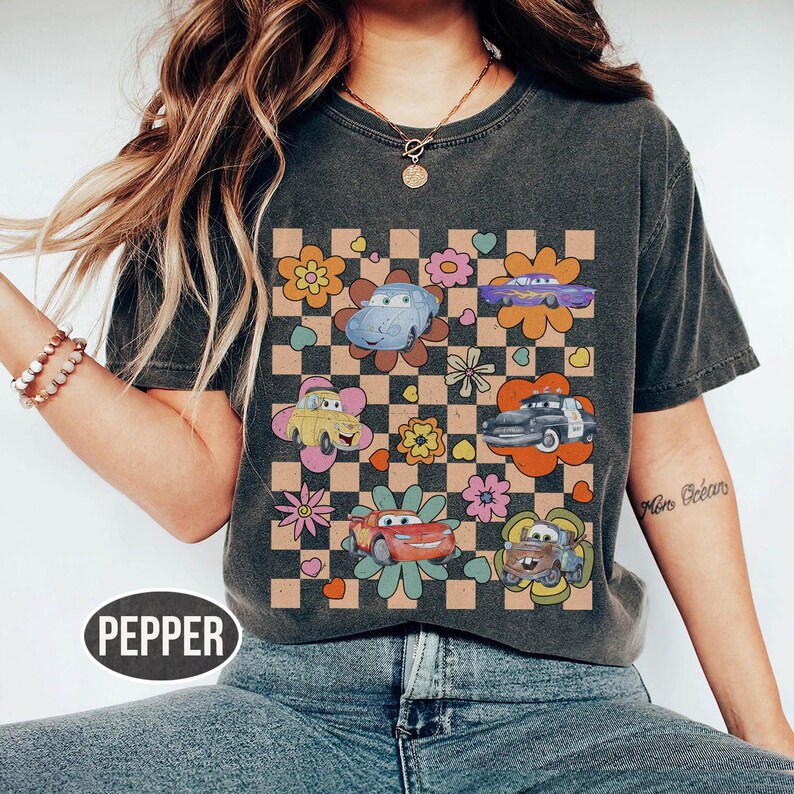 May include: Dark grey t-shirt with a retro design of cartoon cars and flowers on a chequered background. The word "PEPPER" is printed at the bottom. The shirt has short sleeves and a crew neck.