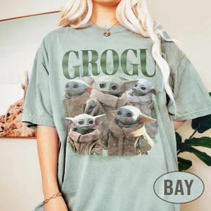 May include: Sage green t-shirt featuring the word "GROGU" in large, green letters above a graphic of Grogu, also known as Baby Yoda, from The Mandalorian. The graphic shows Grogu in various poses. A "BAY" logo is on the bottom right.