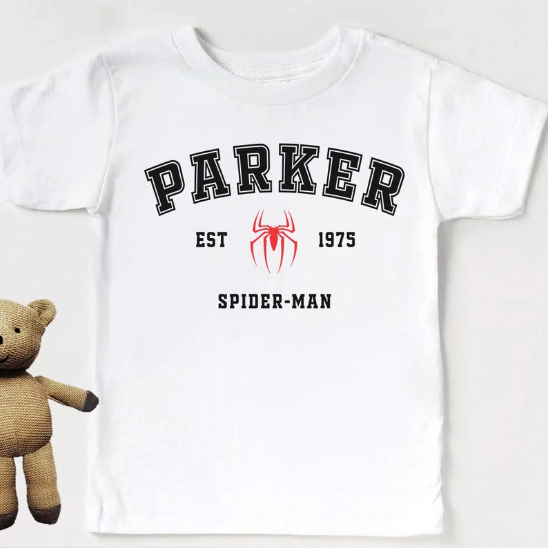 May include: White t-shirt featuring the name "PARKER" in black block letters above "EST 1975" and a red spider emblem. Below is the text "SPIDER-MAN" in black. The shirt is a short-sleeved crew neck.