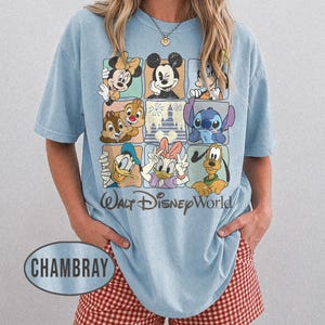 Op de afbeelding: Lichtblauw T-shirt met een raster van Disney-personages, waaronder Mickey Mouse, Minnie Mouse en Donald Duck. De woorden "Walt Disney World" staan eronder gedrukt. Het shirt heeft de kleur chambray.