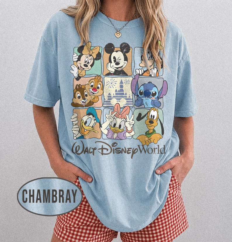 Op de afbeelding: Lichtblauw T-shirt met een raster van Disney-personages, waaronder Mickey Mouse, Minnie Mouse en Donald Duck. De woorden "Walt Disney World" staan eronder gedrukt. Het shirt heeft de kleur chambray.