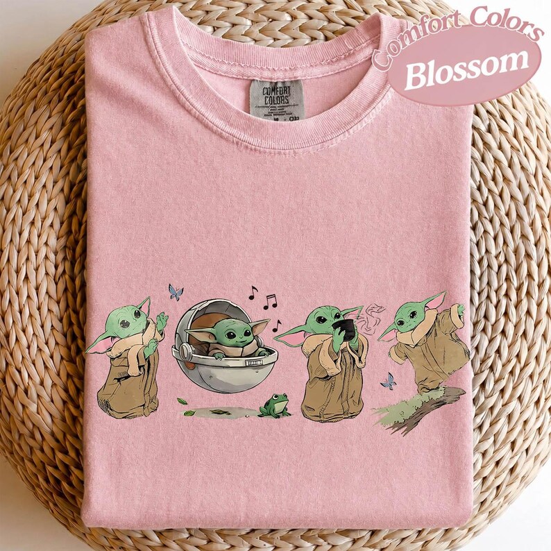 May include: A pale pink Comfort Colors t-shirt with a cartoon illustration of Grogu in different poses. The design includes musical notes, butterflies, and a frog. The shirt has the text "Comfort Colors Blossom".