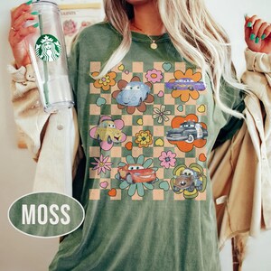 May include: A moss green t-shirt with a retro design featuring cartoon cars and flowers on a checkered background. The cars are in various colours, and the flowers are in shades of yellow, pink, and orange. The shirt has a relaxed fit.