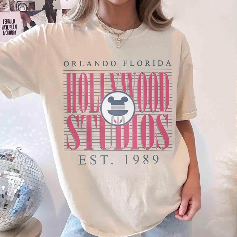 May include: A cream-coloured t-shirt with a pink and grey graphic that reads "Hollywood Studios" with a Mickey Mouse head logo, and "Orlando Florida" above. The text "Est. 1989" is below the graphic. The shirt has short sleeves.