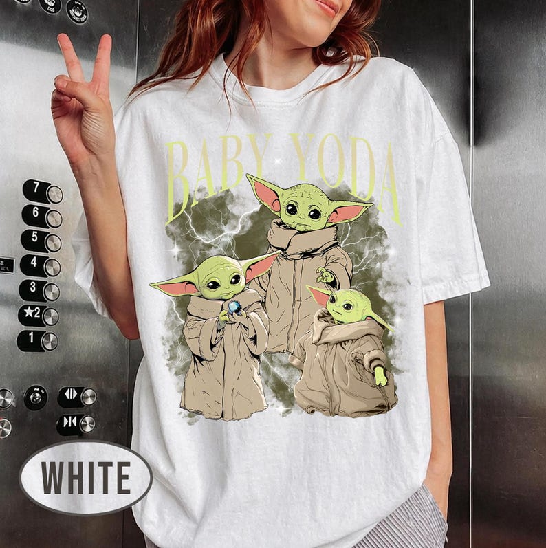 May include: A white short-sleeve t-shirt featuring a graphic print of three green creatures with large ears and tan robes. The text "BABY YODA" is printed in yellow above the characters, set against a dark, smoky background with lightning effects.