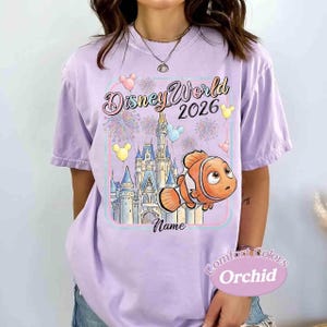 May include: A light purple t-shirt with a Disney World 2026 graphic. The design includes a castle, Nemo the clownfish, fireworks, and balloons. The shirt also has the word "Name" and "Comfort Colors Orchid" printed on it.