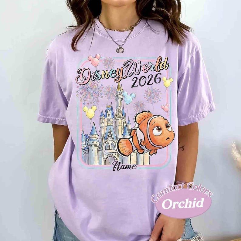 May include: A light purple t-shirt with a Disney World 2026 graphic. The design includes a castle, Nemo the clownfish, fireworks, and balloons. The shirt also has the word "Name" and "Comfort Colors Orchid" printed on it.