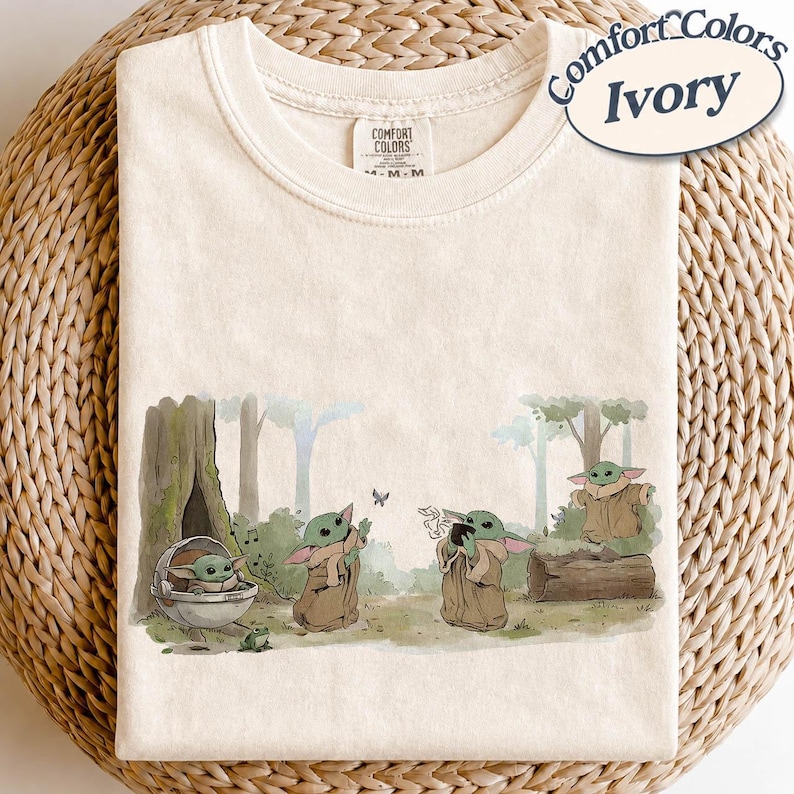 K&ouml;nnte beinhalten: Elfenbeinfarbenes T-Shirt mit einer Grafik von Grogu aus The Mandalorian. Die Illustration zeigt Grogu in verschiedenen Posen in einer Waldlandschaft. Das Shirt tr&auml;gt die Aufschrift "Comfort Colors" und "Ivory".