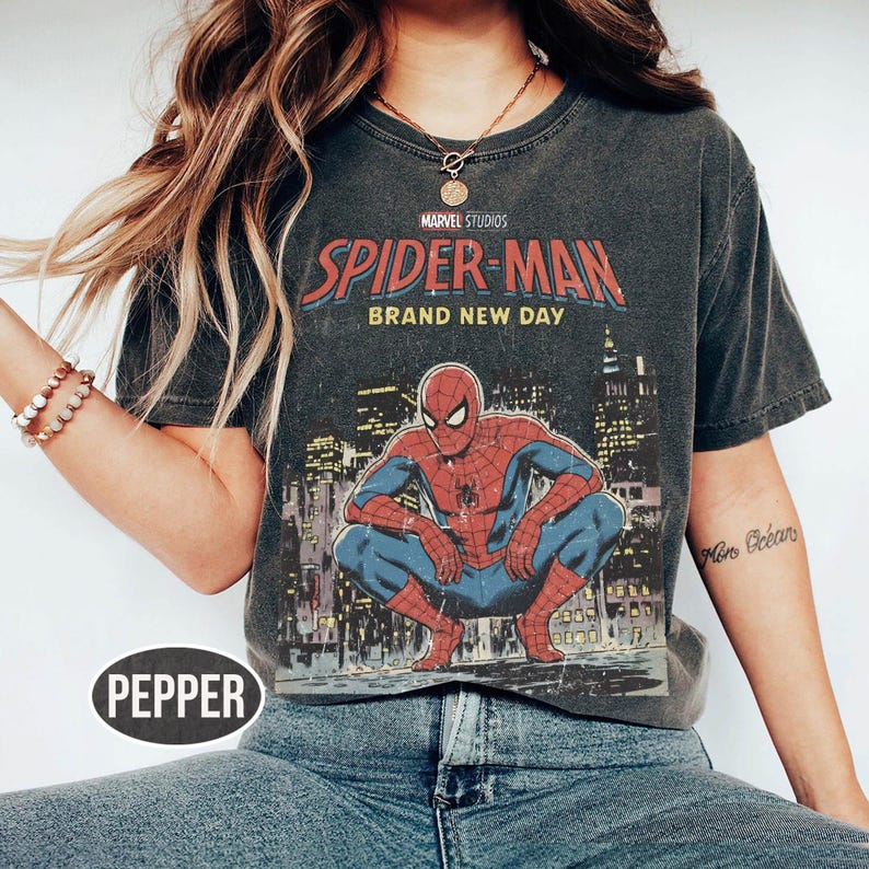 Może przedstawiać: Ciemnoszara koszulka z grafiką Spider-Mana w pozycji kucznej na tle panoramy miasta. Na koszulce widnieje napis "MARVEL STUDIOS SPIDER-MAN BRAND NEW DAY" w kolorze czerwonym i niebieskim.