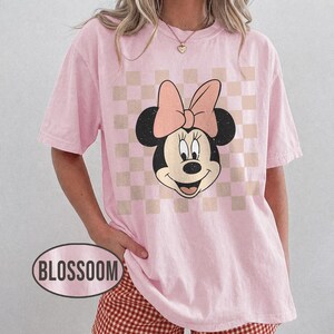 May include: Pink t-shirt featuring a Minnie Mouse graphic with a pink bow and a checkered background. The shirt has short sleeves and a relaxed fit. The word "BLOSSOM" is printed on the bottom left of the shirt.