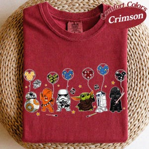 May include: Crimson Comfort Colors t-shirt featuring Star Wars characters and Mickey Mouse-shaped balloons. The design includes BB-8, Chewbacca, a Stormtrooper, Grogu, R2-D2, and Darth Vader. The shirt has a crew neck and is laid flat.
