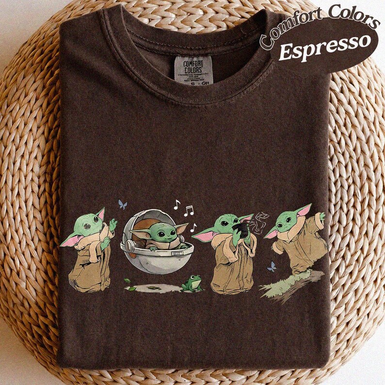 May include: Espresso-coloured t-shirt with a cartoon design of a character from a popular science fiction series. The graphic shows the character in various poses, with musical notes and a frog. The shirt has the Comfort Colors brand label.