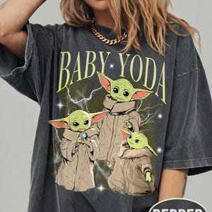 May include: A charcoal gray short-sleeve t-shirt featuring a graphic print of three green creatures in tan robes. The text BABY YODA is printed in light green above the characters, with white lightning bolts and small star accents in the background.