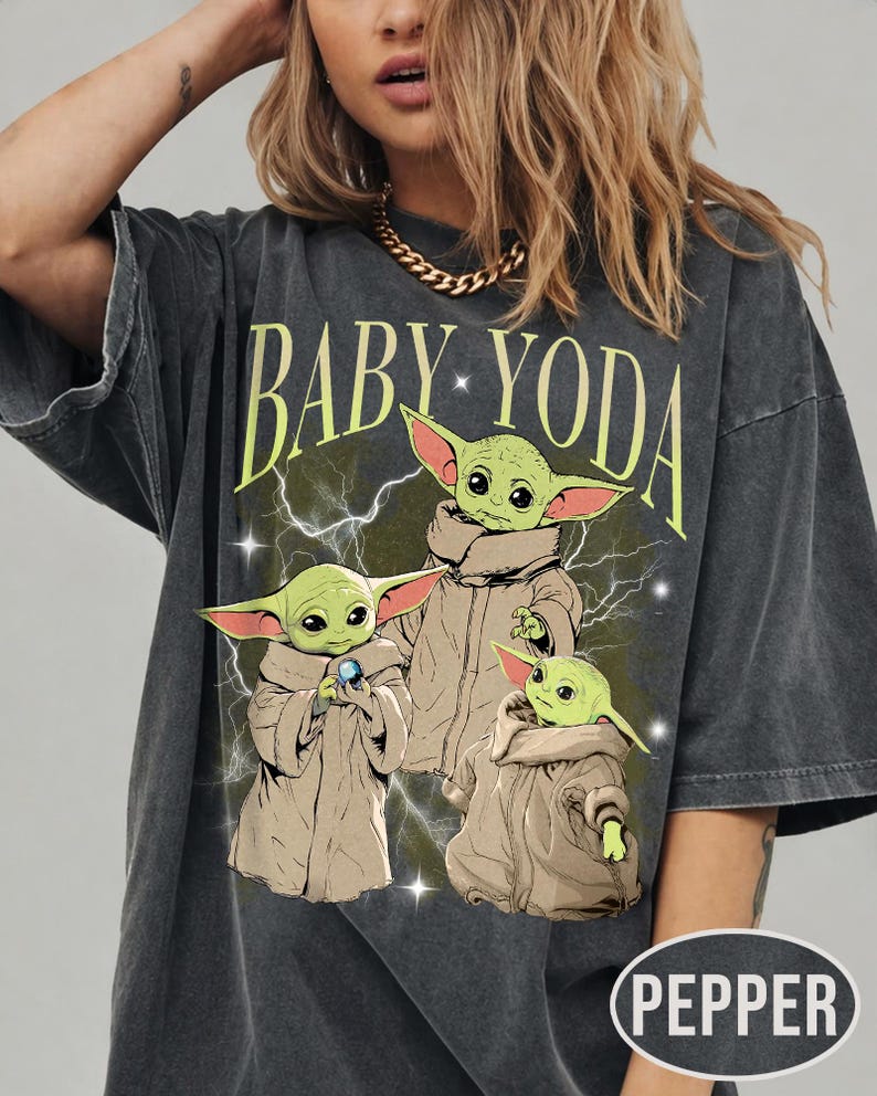 May include: A charcoal gray short-sleeve t-shirt featuring a graphic print of three green creatures in tan robes. The text BABY YODA is printed in light green above the characters, with white lightning bolts and small star accents in the background.