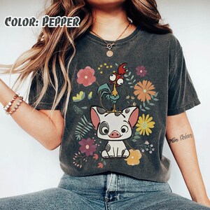 Puede incluir: Camiseta gris oscuro con un dise&ntilde;o de cerdo y gallo de dibujos animados, rodeada de flores coloridas. La camiseta tiene un aspecto vintage y el texto "Color: Pepper" es visible en la esquina superior izquierda.