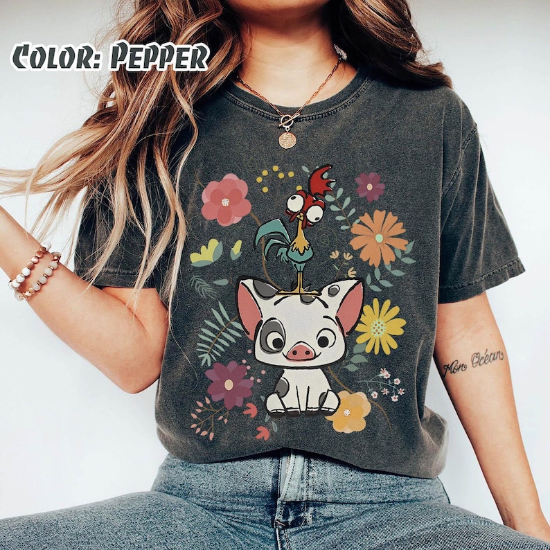 Puede incluir: Camiseta gris oscuro con un dise&ntilde;o de cerdo y gallo de dibujos animados, rodeada de flores coloridas. La camiseta tiene un aspecto vintage y el texto "Color: Pepper" es visible en la esquina superior izquierda.