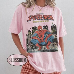 Może przedstawiać: Jasnor&oacute;żowa koszulka z grafiką Spider-Mana z Marvel Studios. Grafika przedstawia Spider-Mana kucającego przed panoramą miasta, z napisem "Spider-Man Brand New Day". Koszulka ma kr&oacute;tkie rękawy.