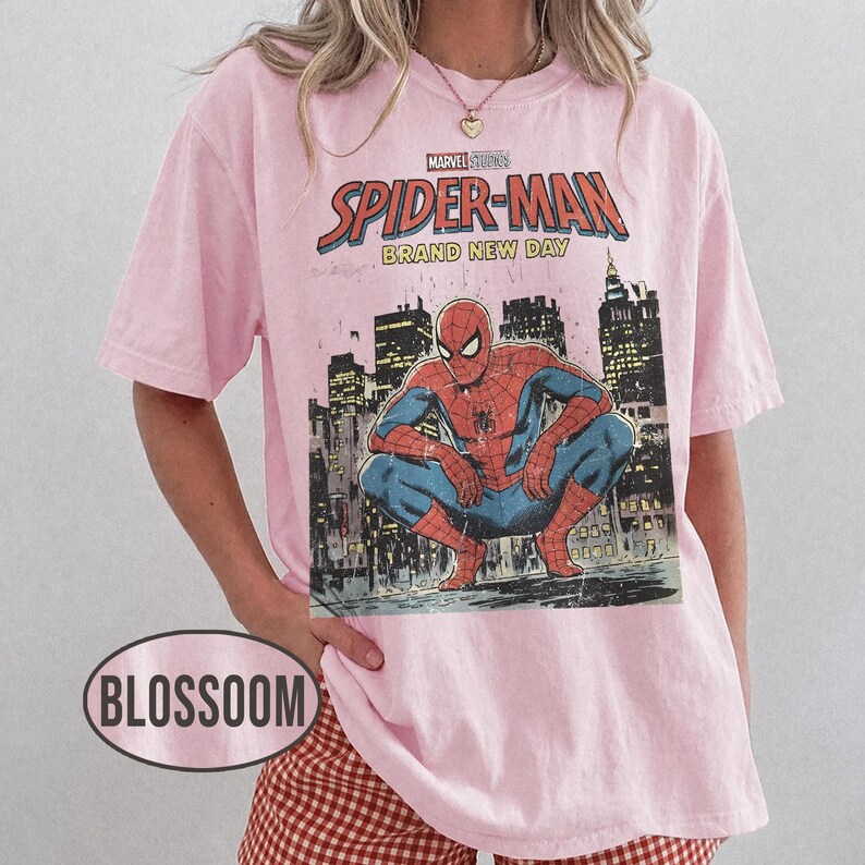 Może przedstawiać: Jasnor&oacute;żowa koszulka z grafiką Spider-Mana z Marvel Studios. Grafika przedstawia Spider-Mana kucającego przed panoramą miasta, z napisem "Spider-Man Brand New Day". Koszulka ma kr&oacute;tkie rękawy.