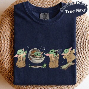 May include: True Navy Comfort Colors t-shirt with a graphic of a character from a popular science fiction series. The design includes the character in various poses, with musical notes and a small frog. The shirt is on a woven surface.