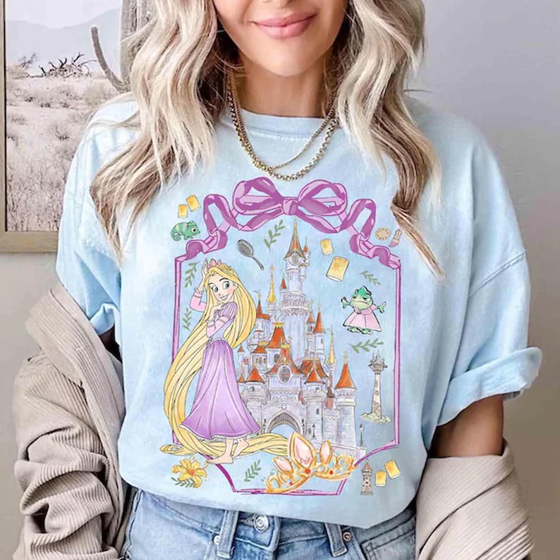May include: Light blue t-shirt featuring a cartoon illustration of a princess with long blonde hair, a castle, and other fairytale elements. The design is framed by a pink ribbon and includes a crown and a frog.
