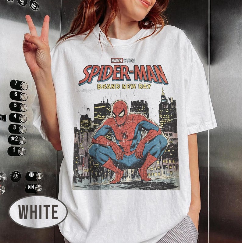 Może przedstawiać: Biała koszulka z grafiką Spider-Mana z Marvel Studios. Projekt zawiera napis "SPIDER-MAN BRAND NEW DAY" oraz ilustrację Spider-Mana w kucnięciu na tle panoramy miasta. Odzież codzienna.