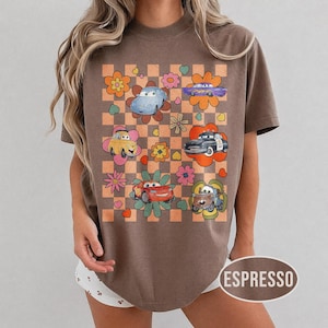 May include: Espresso-coloured t-shirt with a retro design featuring cartoon cars and flowers on a chequered background. The cars are in various colours, and the flowers are in shades of orange, pink, and yellow. The shirt has a relaxed fit.