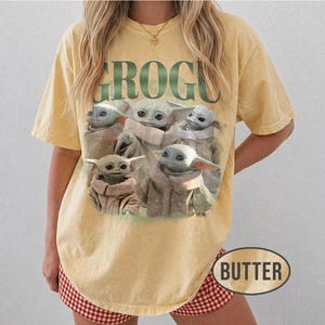 May include: Butter-colored t-shirt featuring a graphic of Grogu, also known as Baby Yoda, with the word "GROGU" in green block letters. The image shows multiple Grogu characters in tan robes. The shirt has short sleeves.