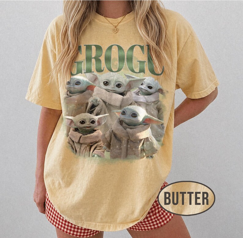 May include: Butter-colored t-shirt featuring a graphic of Grogu, also known as Baby Yoda, with the word "GROGU" in green block letters. The image shows multiple Grogu characters in tan robes. The shirt has short sleeves.