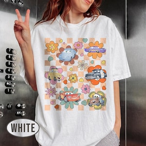 May include: White t-shirt with a vibrant graphic of cartoon cars and flowers. The design showcases different car characters on a checkered background with floral details. The shirt has a loose fit.