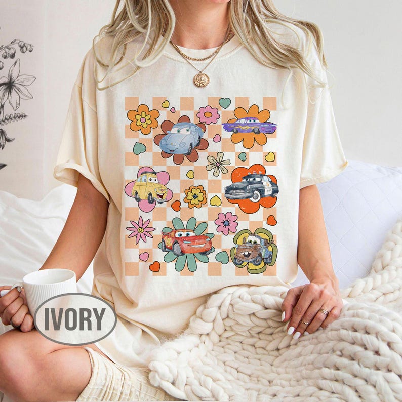 May include: Cream-coloured t-shirt with a colourful graphic of cartoon cars and flowers on a checkered background. The word "IVORY" is printed on a white mug. The design includes various car characters in a retro style.