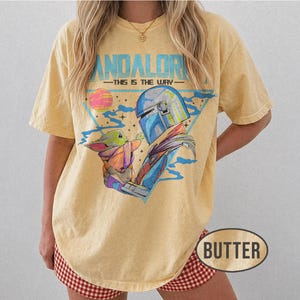 May include: Butter-coloured t-shirt featuring a graphic of The Mandalorian and Grogu, with the text "MANDALORIAN - THIS IS THE WAY." The design includes a blue helmet, a pink and yellow planet, and a triangle. The shirt has short sleeves.
