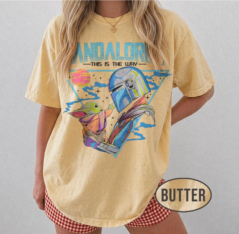 May include: Butter-coloured t-shirt featuring a graphic of The Mandalorian and Grogu, with the text "MANDALORIAN - THIS IS THE WAY." The design includes a blue helmet, a pink and yellow planet, and a triangle. The shirt has short sleeves.