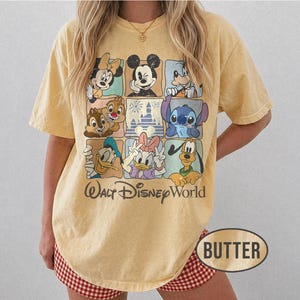 Op de afbeelding: Boterkleurig T-shirt met een raster van Disney-personages, waaronder Mickey Mouse, Minnie Mouse, Donald Duck en Stitch. De woorden "Walt Disney World" staan eronder gedrukt. Het shirt heeft korte mouwen en een relaxte pasvorm.