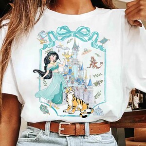 May include: White t-shirt featuring a cartoon illustration of a princess in a teal dress, a tiger, and a castle. The design includes a teal bow and decorative elements. The shirt has a brown belt and blue jeans.