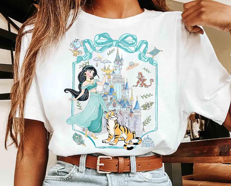 May include: White t-shirt featuring a cartoon illustration of a princess in a teal dress, a tiger, and a castle. The design includes a teal bow and decorative elements. The shirt has a brown belt and blue jeans.