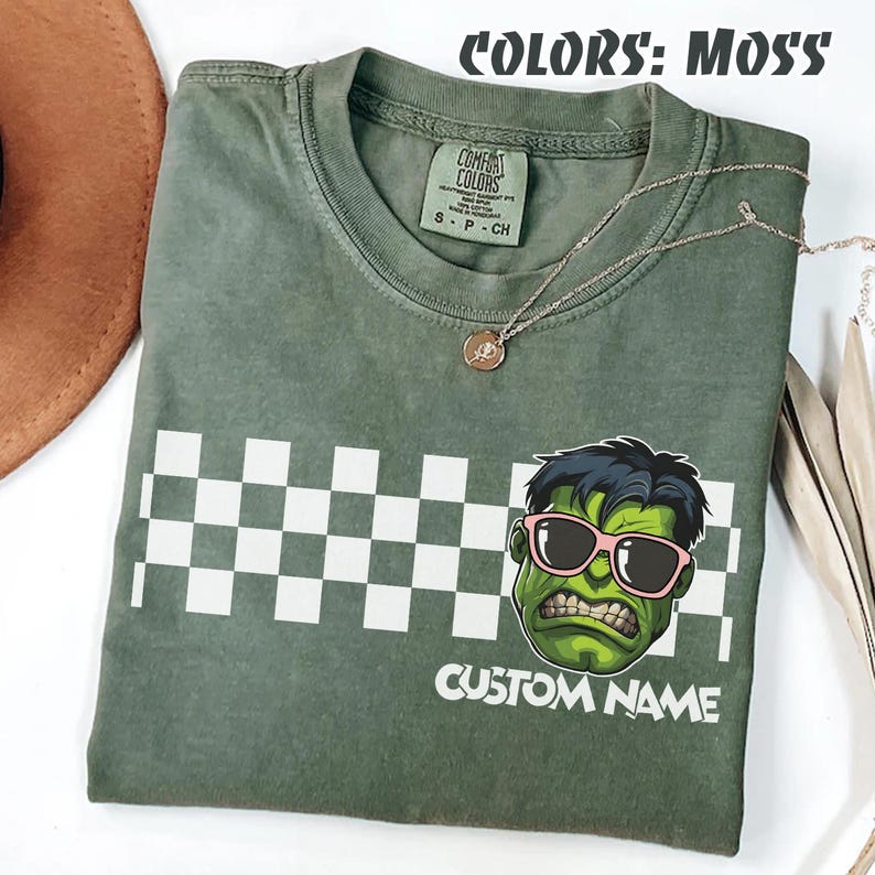 May include: Moss green t-shirt with a graphic of a green cartoon character wearing pink sunglasses. The shirt has a white checkered pattern and the words "CUSTOM NAME" printed below the graphic. A gold necklace is draped across the shirt.