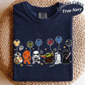 May include: True navy blue t-shirt featuring a graphic of Star Wars characters and Mickey Mouse balloons. The design includes BB-8, Chewbacca, a Stormtrooper, Grogu, R2-D2, and Darth Vader. The shirt is made by Comfort Colors.