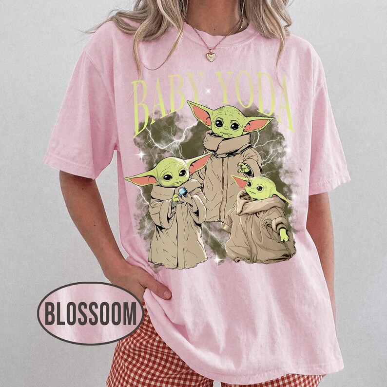 May include: A light pink short-sleeve t-shirt featuring a graphic print of three green creatures in tan robes with the text BABY YODA in yellow letters above them. The design includes a dark, smoky background with small white star accents.