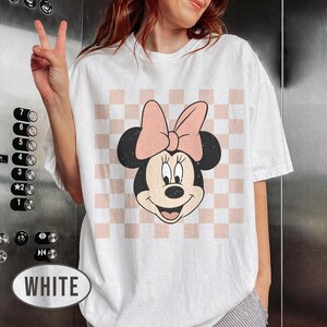 May include: White t-shirt featuring a Minnie Mouse graphic. The design includes Minnie's face with a pink bow, set against a pink and white checkered background. The shirt is a casual, oversized fit.