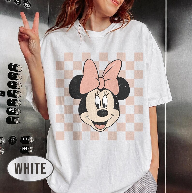 May include: White t-shirt featuring a Minnie Mouse graphic. The design includes Minnie's face with a pink bow, set against a pink and white checkered background. The shirt is a casual, oversized fit.
