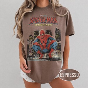 Może przedstawiać: Koszulka w kolorze espresso z grafiką Spider-Mana w stylu vintage. Projekt zawiera napis "Marvel Studios Spider-Man Brand New Day" nad ilustracją Spider-Mana kucającego przed panoramą miasta.
