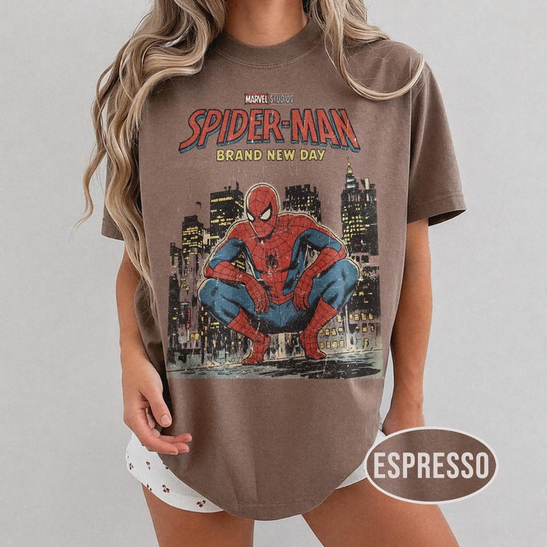 Może przedstawiać: Koszulka w kolorze espresso z grafiką Spider-Mana w stylu vintage. Projekt zawiera napis "Marvel Studios Spider-Man Brand New Day" nad ilustracją Spider-Mana kucającego przed panoramą miasta.