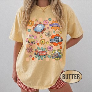 May include: A butter-yellow t-shirt with a retro design. It features cartoon cars and floral elements set against a checkered background with flowers and hearts. The word "BUTTER" is printed at the bottom.