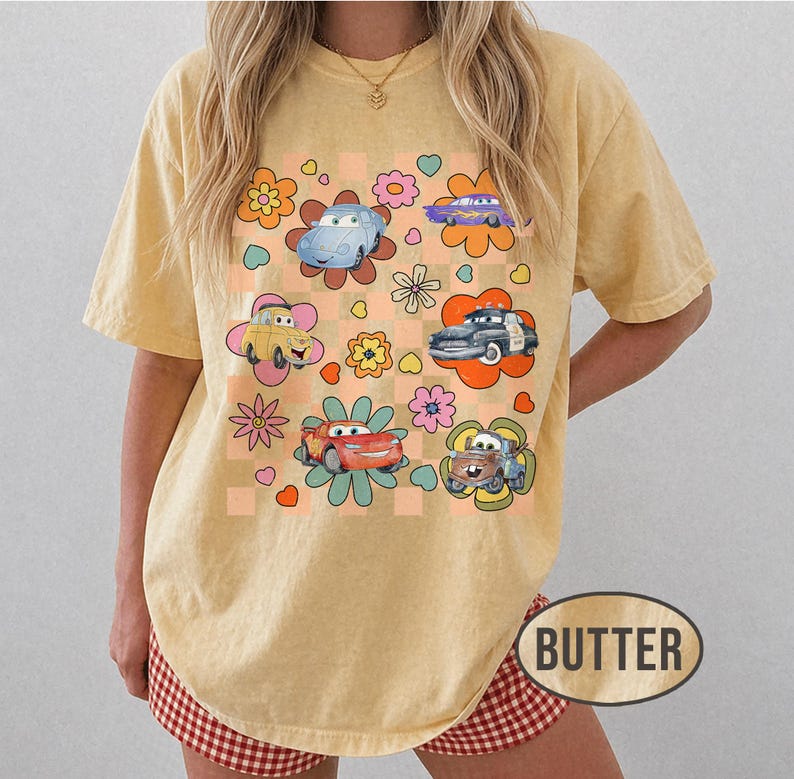 May include: A butter-yellow t-shirt with a retro design. It features cartoon cars and floral elements set against a checkered background with flowers and hearts. The word "BUTTER" is printed at the bottom.