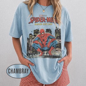 Może przedstawiać: Jasnoniebieska koszulka z grafiką Spider-Mana w kucającej pozie na tle panoramy miasta. Koszulka ma nadruk "Marvel Studios Spider-Man Brand New Day". Modelka ma na sobie czerwono-białe szorty w kratę. Styl casual.