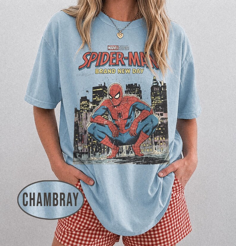 Może przedstawiać: Jasnoniebieska koszulka z grafiką Spider-Mana w kucającej pozie na tle panoramy miasta. Koszulka ma nadruk "Marvel Studios Spider-Man Brand New Day". Modelka ma na sobie czerwono-białe szorty w kratę. Styl casual.